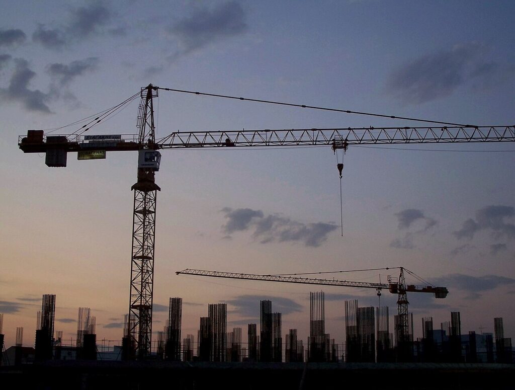 Real documentary photograph of two construction cranes silhouetted at dusk above a worksite in Athens, illustrating the Weekly Lens article about the practical and financial cost of ambitious building plans.